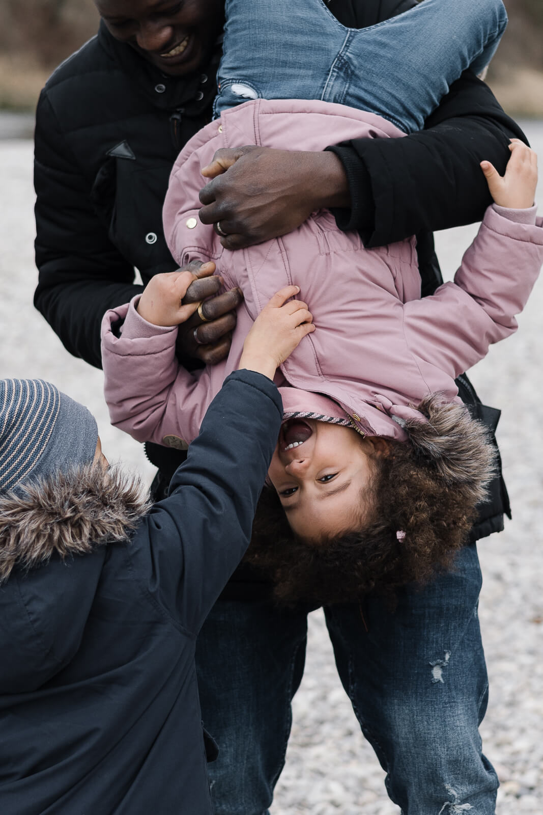 Ein kleiner Junge kitzelt seine Schwester am Bauch während der Papa der Kinder das Mädchen kopfüber im Arm hält